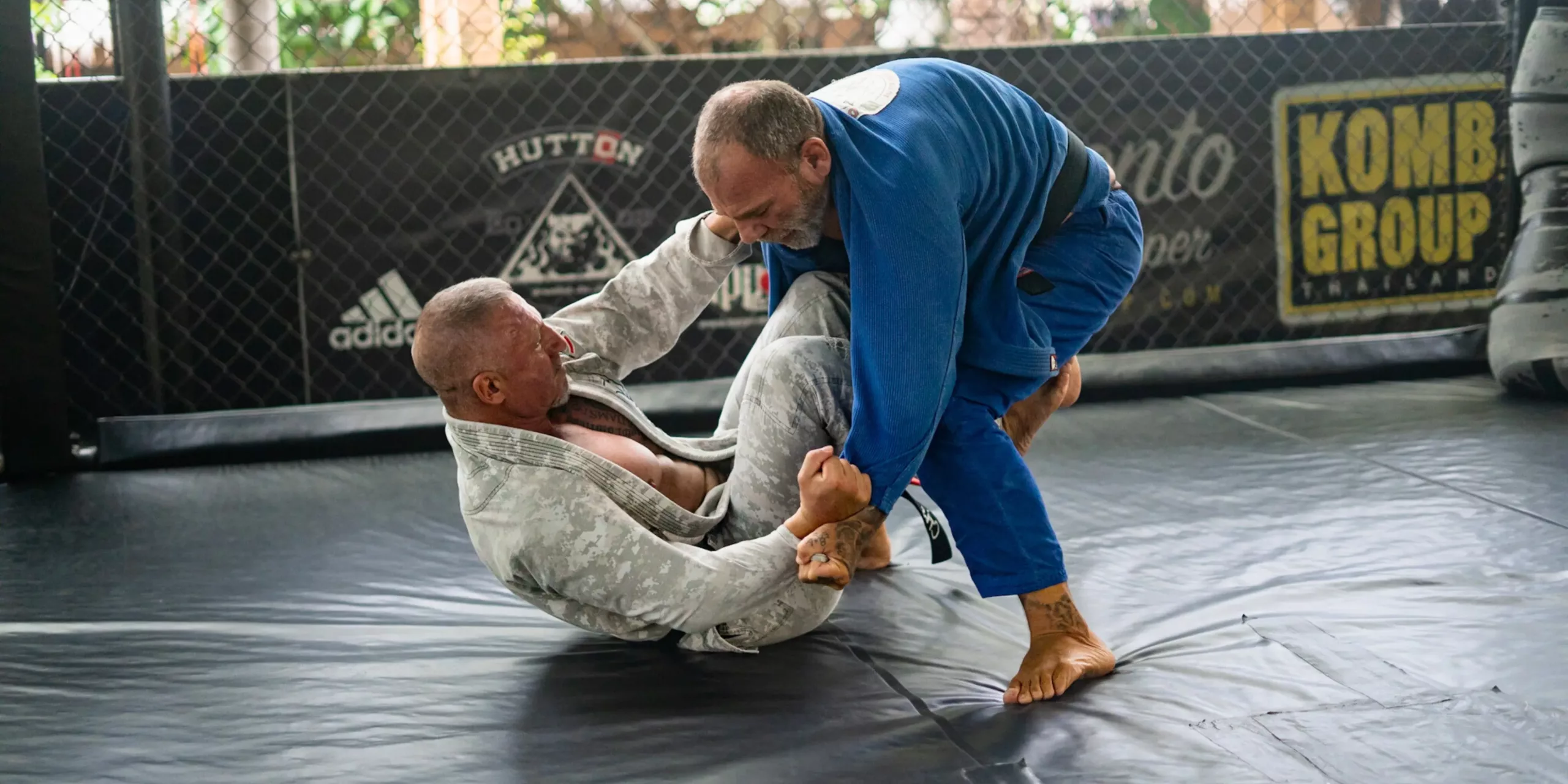 A Brazilian Jiu-Jitsu athlete on the ground works a complex guard against a standing opponent, showcasing the technical and strategic depth of ground combat in BJJ.