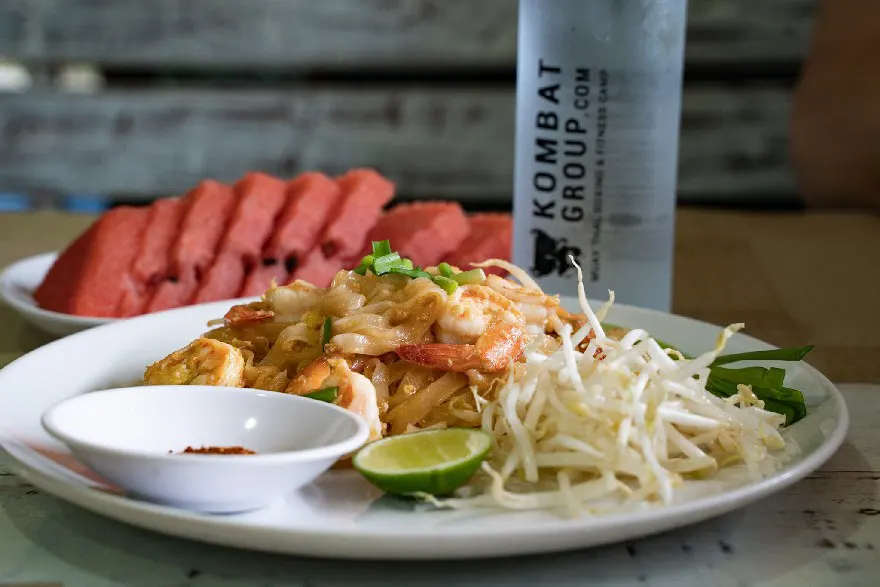 Plate of traditional Pad Thai noodles with shrimp, bean sprouts, and lime, ready to be enjoyed with a side of chili powder and a backdrop of sliced watermelon and a branded water bottle.