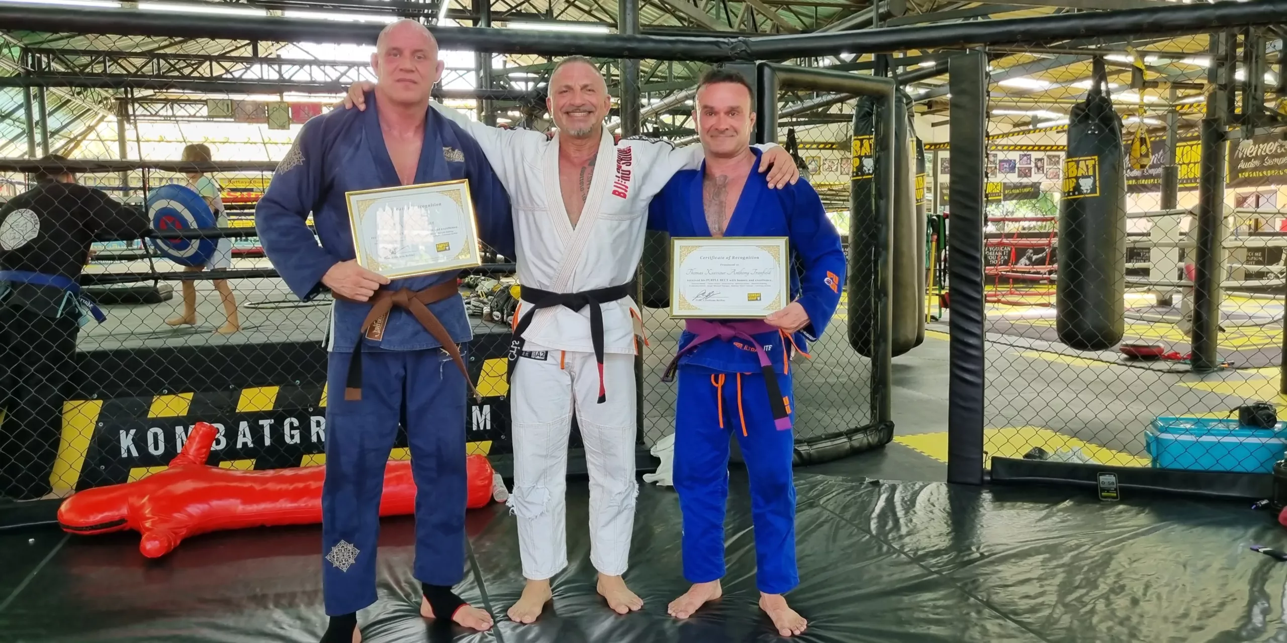Three Brazilian Jiu-Jitsu students smiling proudly in the dojo, holding their promotion certificates and wearing their gis with newly earned belts, celebrating their progress and dedication to the martial art.
