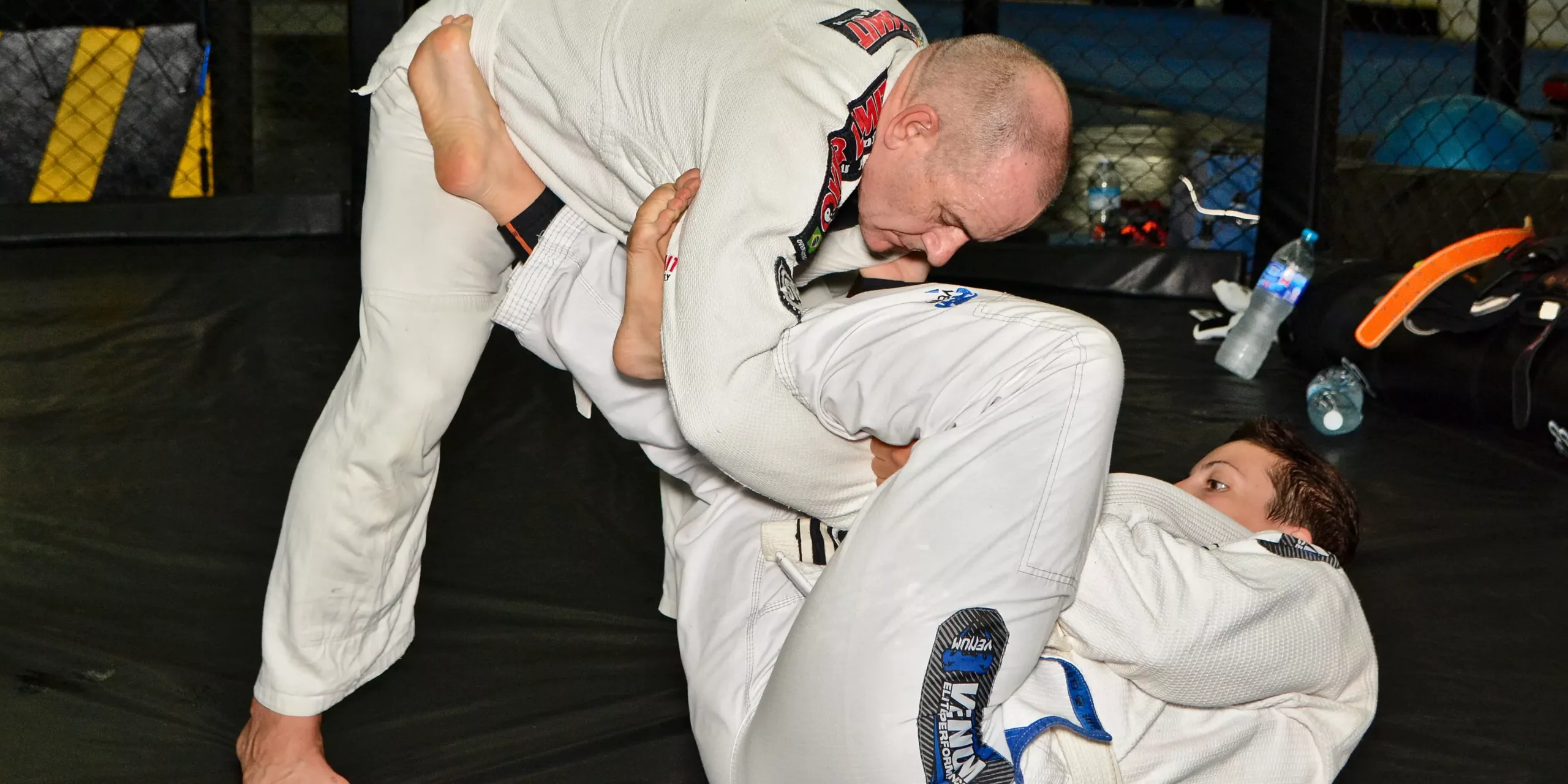 A BJJ practitioner in a white gi works to pass the guard of an opponent, showcasing skill and control in a grappling training session on the mat.