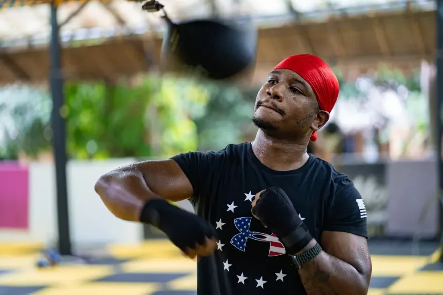 A boxer with a red headband and a star-spangled black shirt practices an uppercut on a speed bag, showcasing concentration and technique in a sunlit gym.