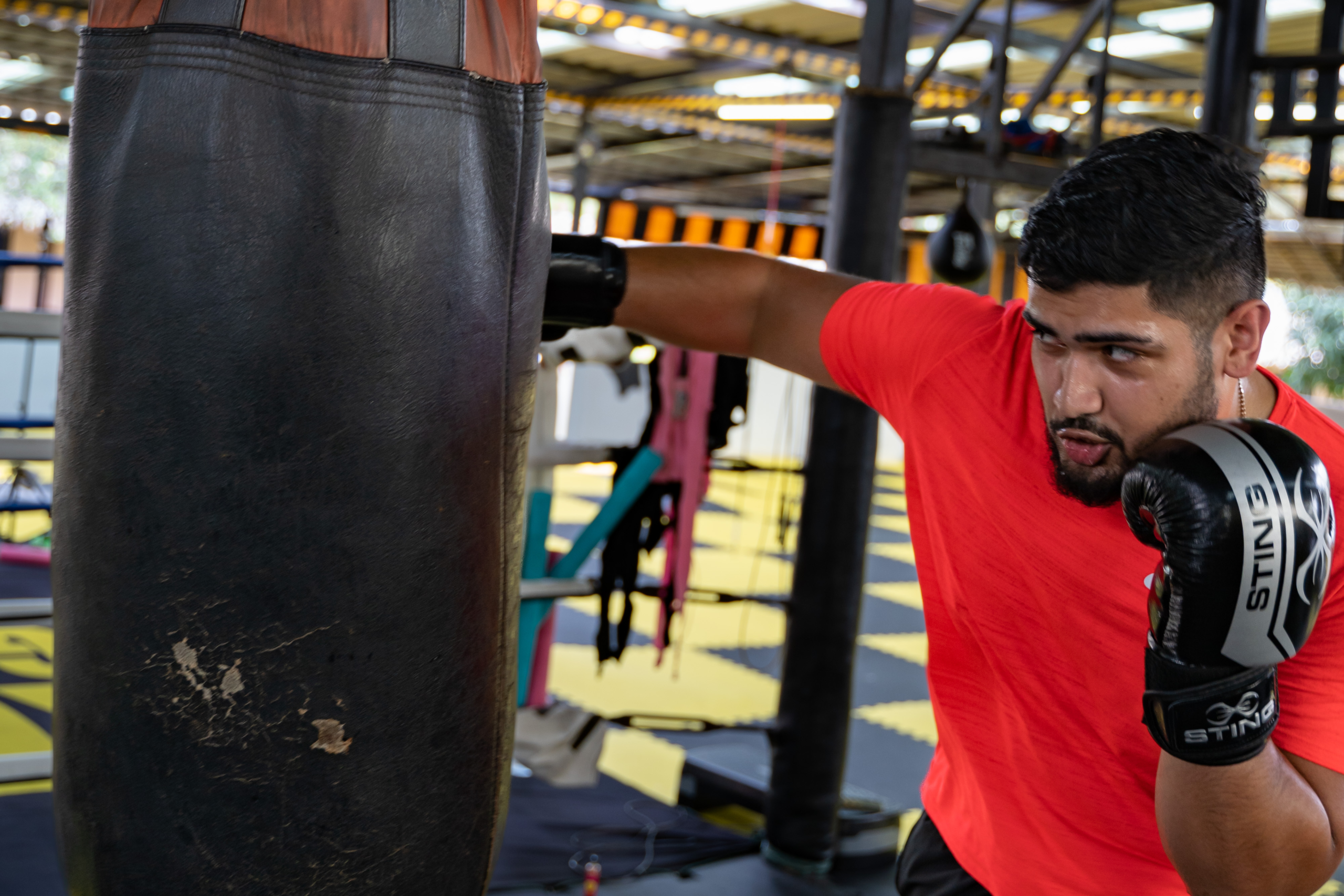 Focused man in a red t-shirt and black boxing gloves practicing a punch on a heavy bag at a self-defense training session, in a gym with visible equipment in the background.