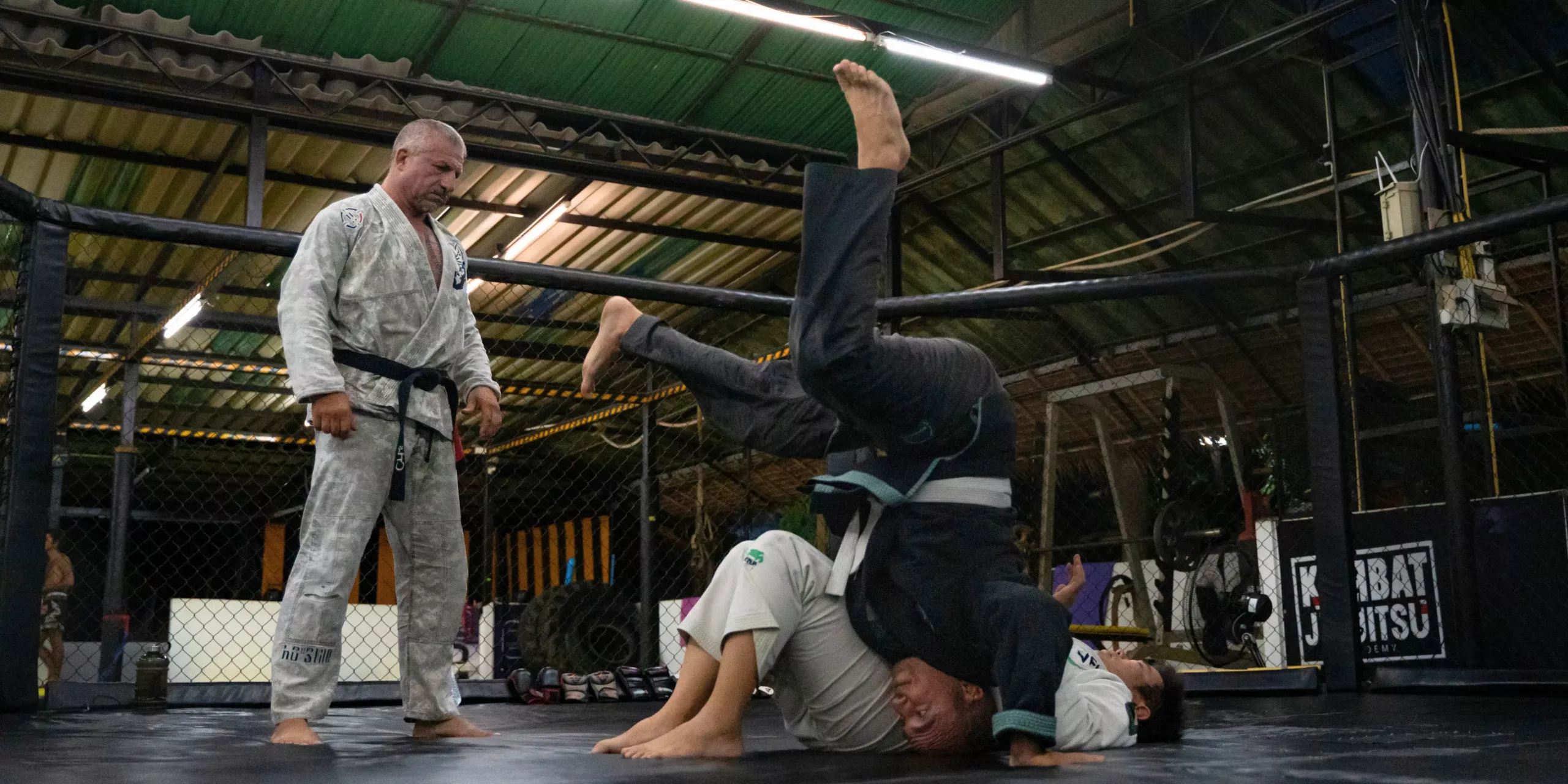A Brazilian Jiu-Jitsu instructor observes two students, one executing an inverted guard maneuver while the other attempts to maintain position, in a dynamic training exercise on the mats.