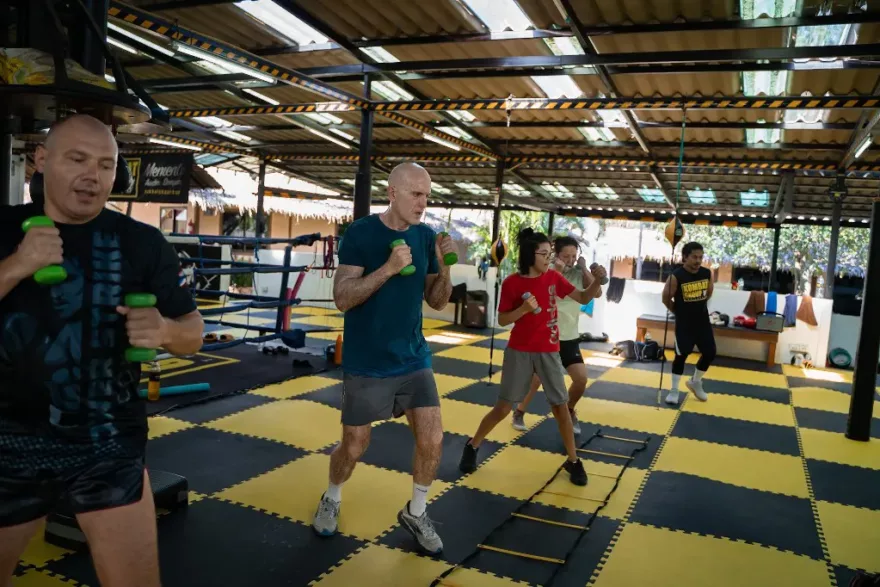 A group of fitness enthusiasts of various ages performing a cardio workout with dumbbells at a martial arts gym, with a focus on movement and agility on yellow and black mats.