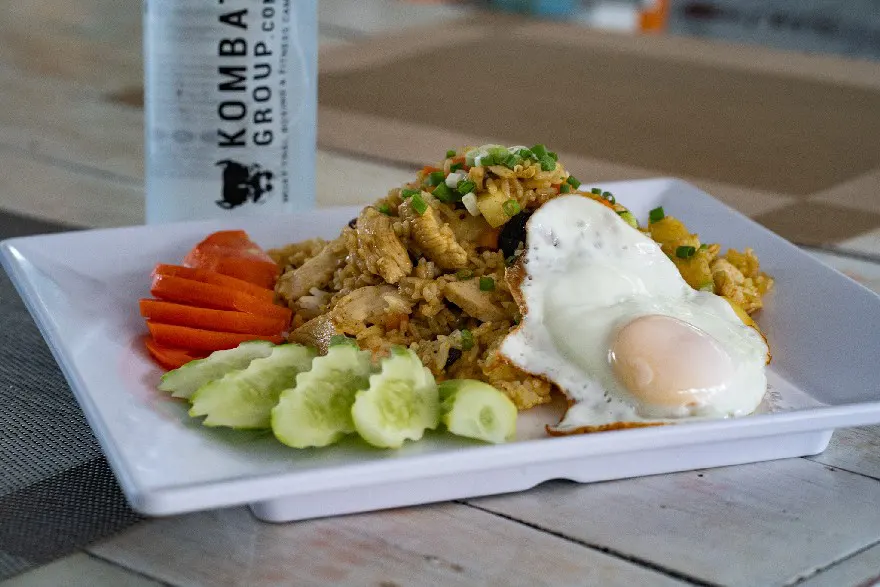 A plate of chicken fried rice topped with a sunny-side-up egg, garnished with scallions, accompanied by slices of cucumber and carrot, with a branded water bottle in the background.