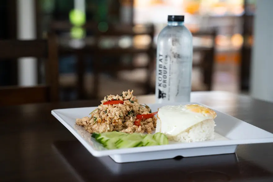 Traditional Laarb chicken topped with a fried egg, served with jasmine rice and cucumber slices, presented on a white plate with a branded water bottle in the background.