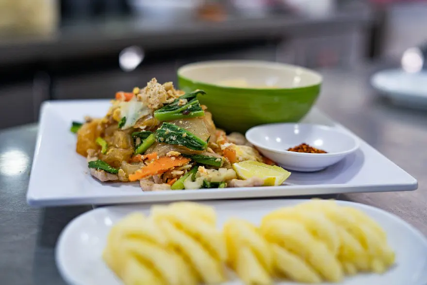 A vibrant dish of stir-fried vegetables topped with crispy tofu, accompanied by a bowl of soup, chili sauce, and fresh slices of pineapple on a sleek kitchen countertop.
