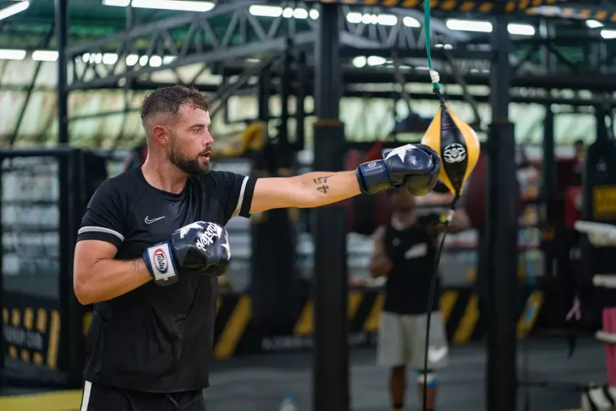 A determined male boxer with a beard, wearing black athletic gear and boxing gloves, extends a powerful jab towards a speed bag in a gym with other athletes training in the background.