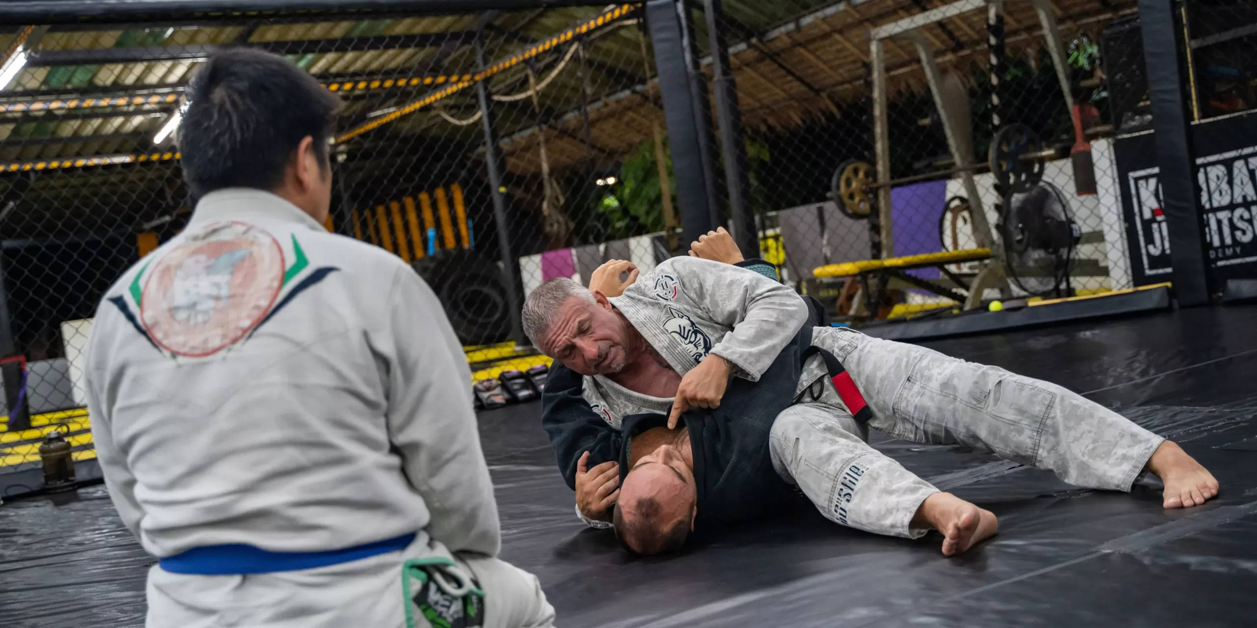 A Brazilian Jiu-Jitsu student attempts to escape a tight side control by an experienced practitioner in a grey gi, while another looks on, in a focused and realistic rolling session on the mats.