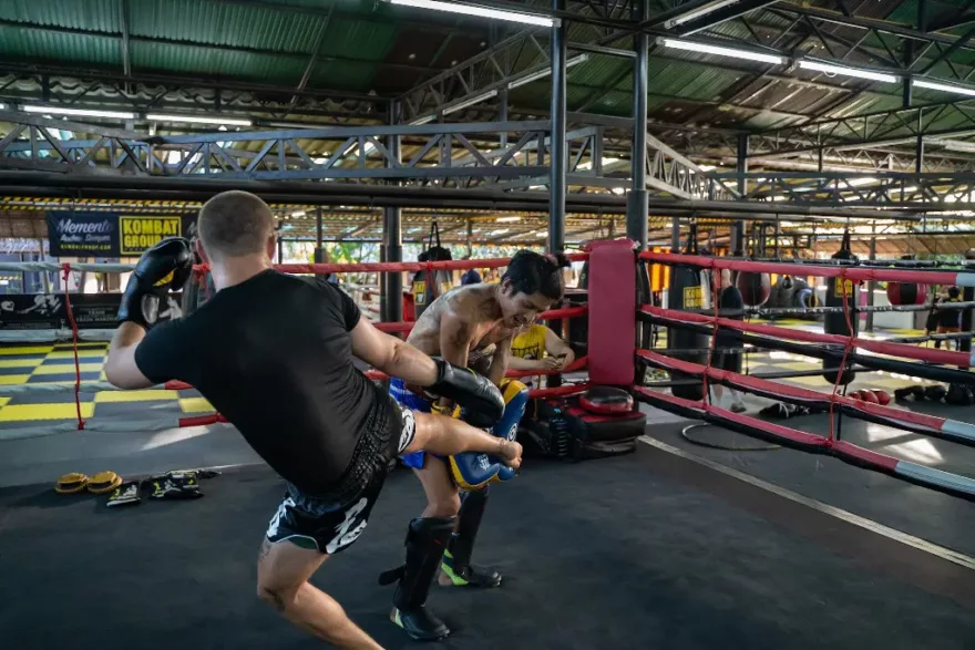 A kickboxer in black shorts delivers a high kick to his sparring partner holding pads, inside a spacious gym with a boxing ring and equipment in the background.