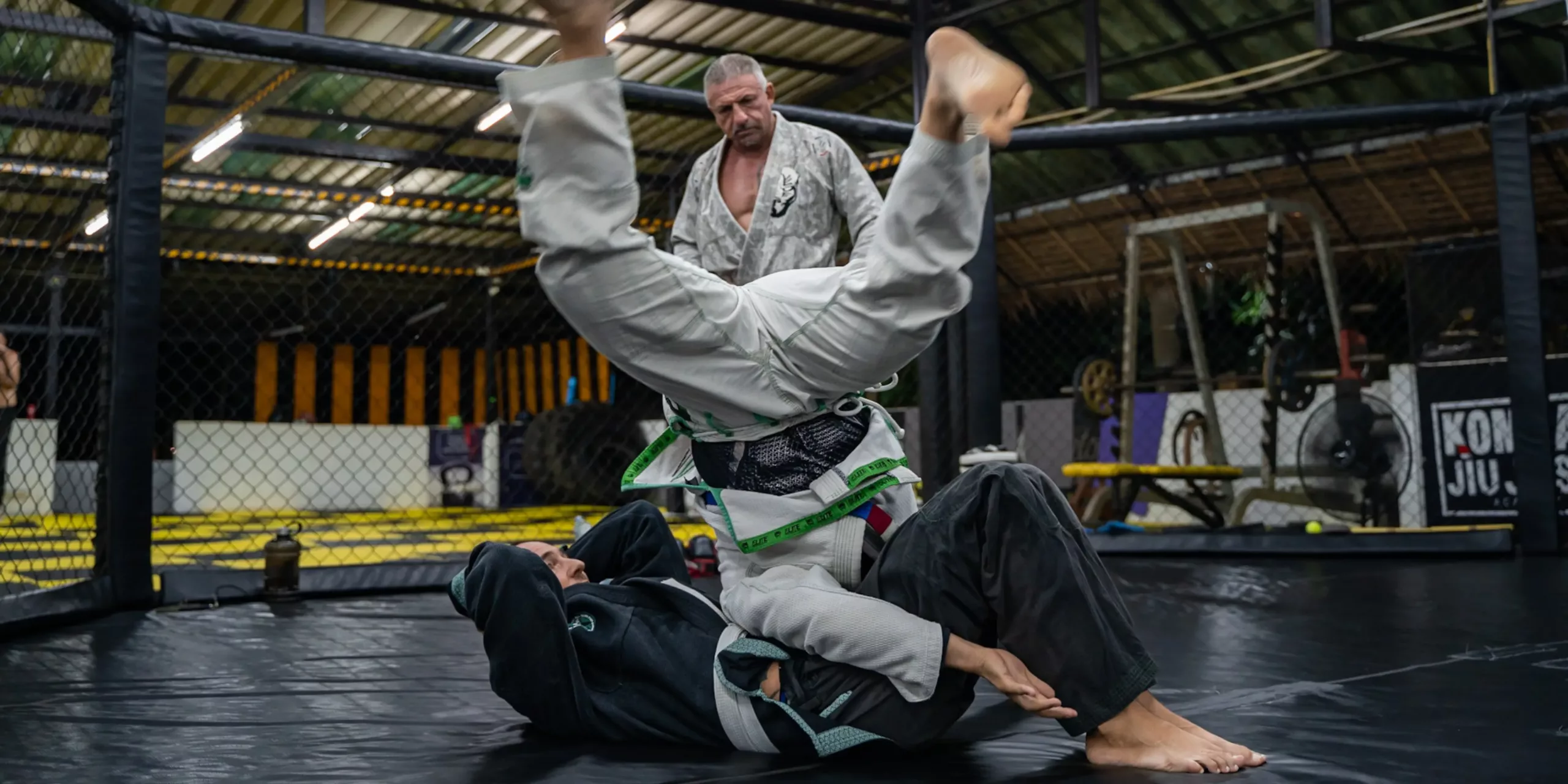 A Brazilian Jiu-Jitsu student in a white gi performs a sweeping technique from the bottom, lifting his opponent into the air, with their instructor supervising the drill in the dojo.
