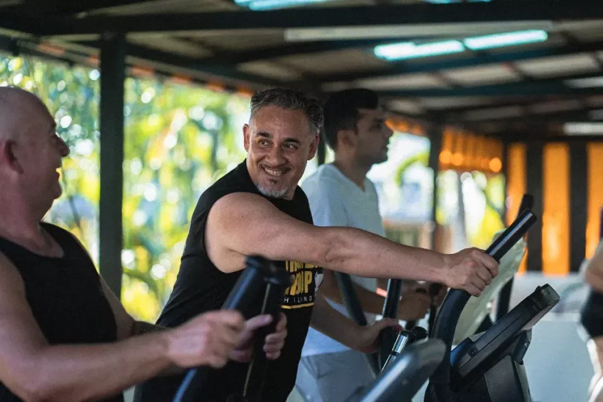 A smiling man in a black sleeveless top engaging with workout companions on elliptical machines at a gym, with lush greenery visible in the background.