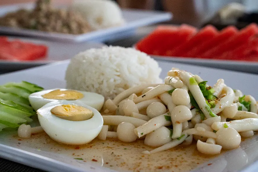 A tantalizing plate showcasing a spicy mushroom salad with sliced hard-boiled eggs, crisp cucumber, and steamed rice, with a slice of watermelon visible in the background.