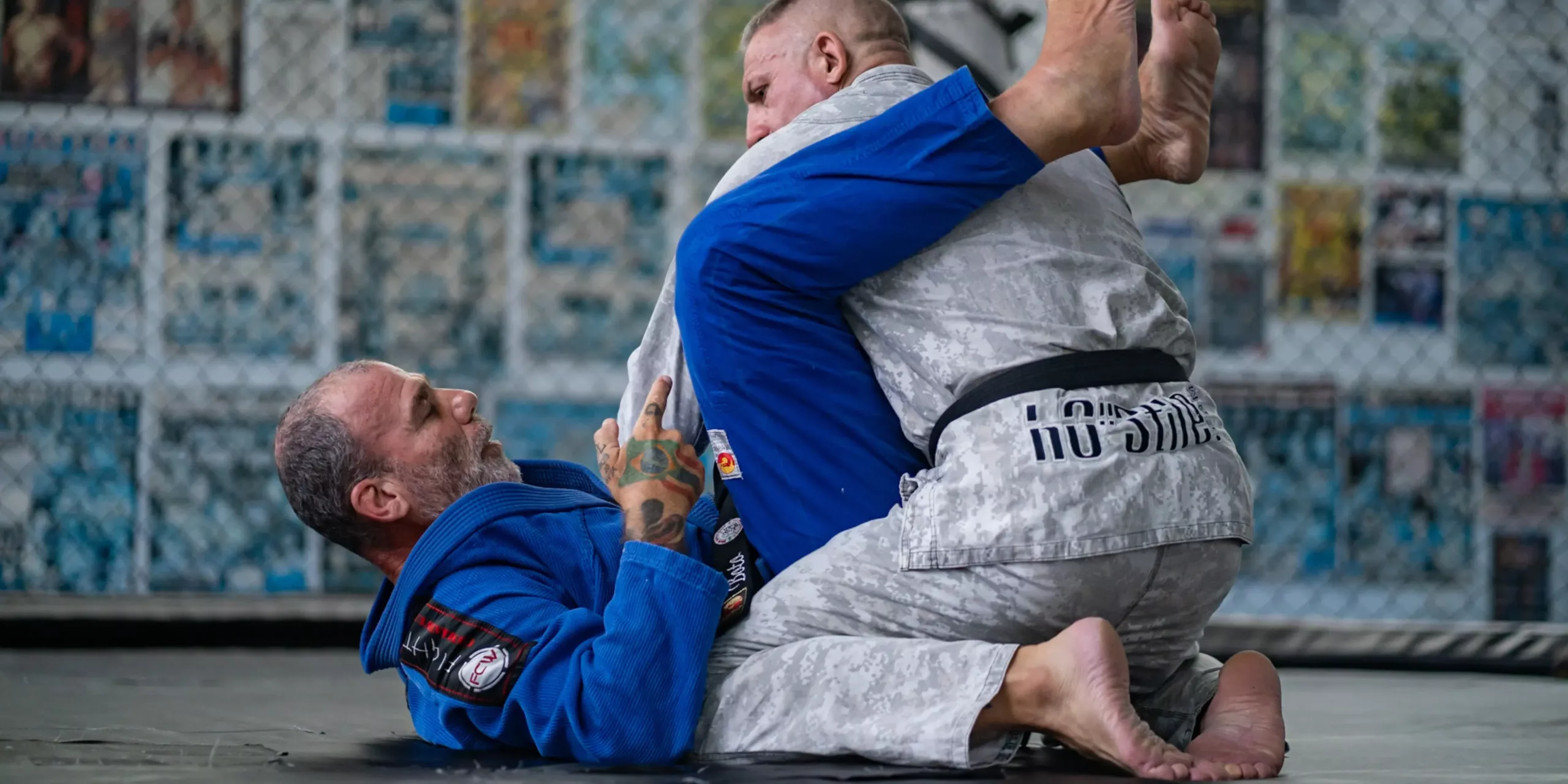 Two Brazilian Jiu-Jitsu practitioners engaged in technical training, with one on his back in guard position while the other works to pass, amidst the backdrop of a dojo with BJJ lineage displays.