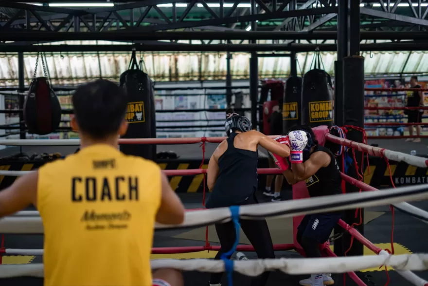 A boxing coach in a yellow shirt oversees two athletes sparring in a boxing ring, with punching bags and gym equipment in the background.