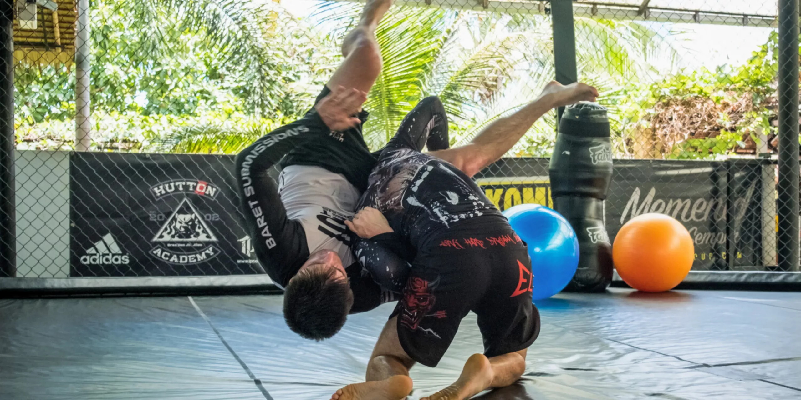 An action-packed moment in a Brazilian Jiu-Jitsu class, where a student in black athletic wear attempts a dynamic guard pass with a flip over his partner, who is on the mat trying to maintain guard.