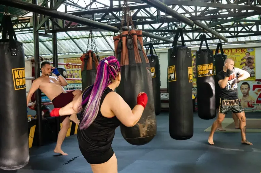 A woman with purple braided hair throws a high kick at a heavy bag while two men practice their boxing skills in the background in a well-equipped Muay Thai gym.