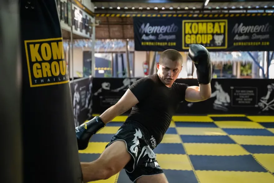 A focused Muay Thai fighter in black attire practicing punches on a heavy bag in a well-equipped gym with Kombat Group branding in the background.