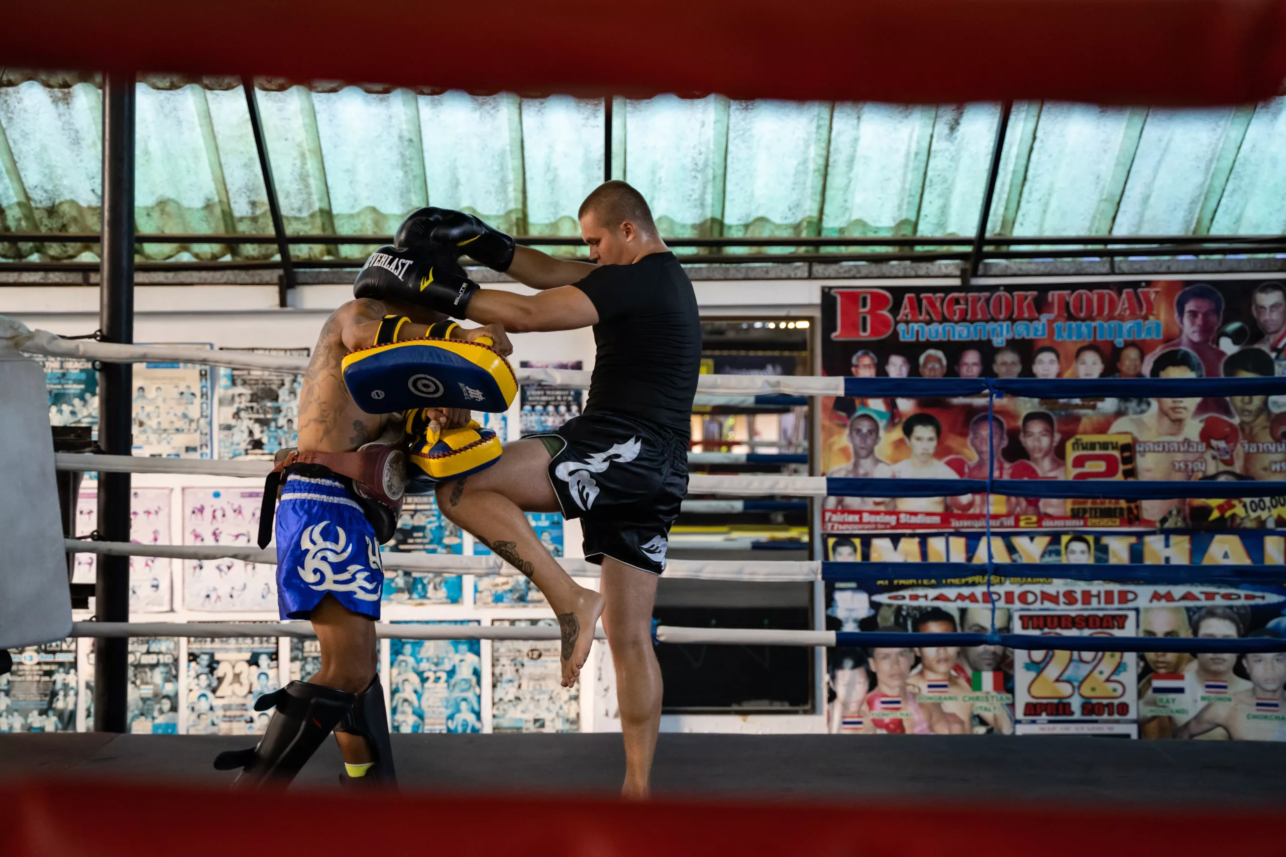 An athlete in black Muay Thai shorts delivers a powerful knee strike to a pad held by a trainer, against the backdrop of a ring and a wall adorned with fight posters.