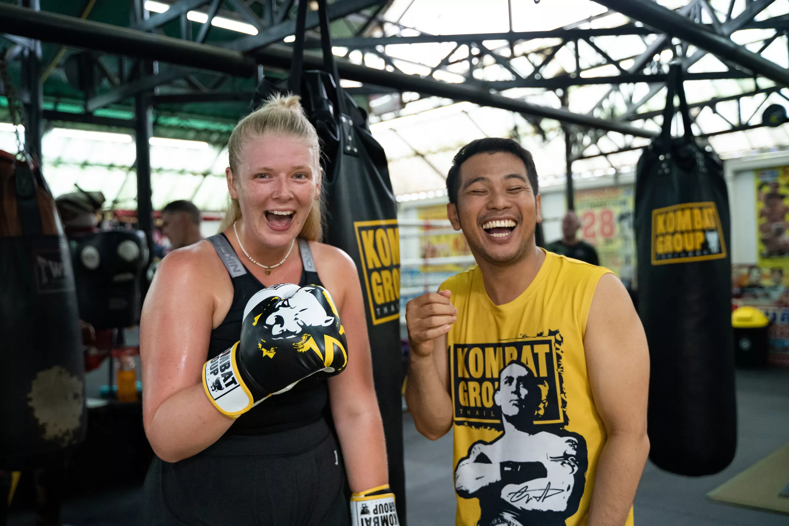 A joyful moment captured between a Muay Thai coach and a student, a woman and a man, laughing together with a sense of accomplishment and camaraderie after a training session, in a gym surrounded by equipment and punching bags.