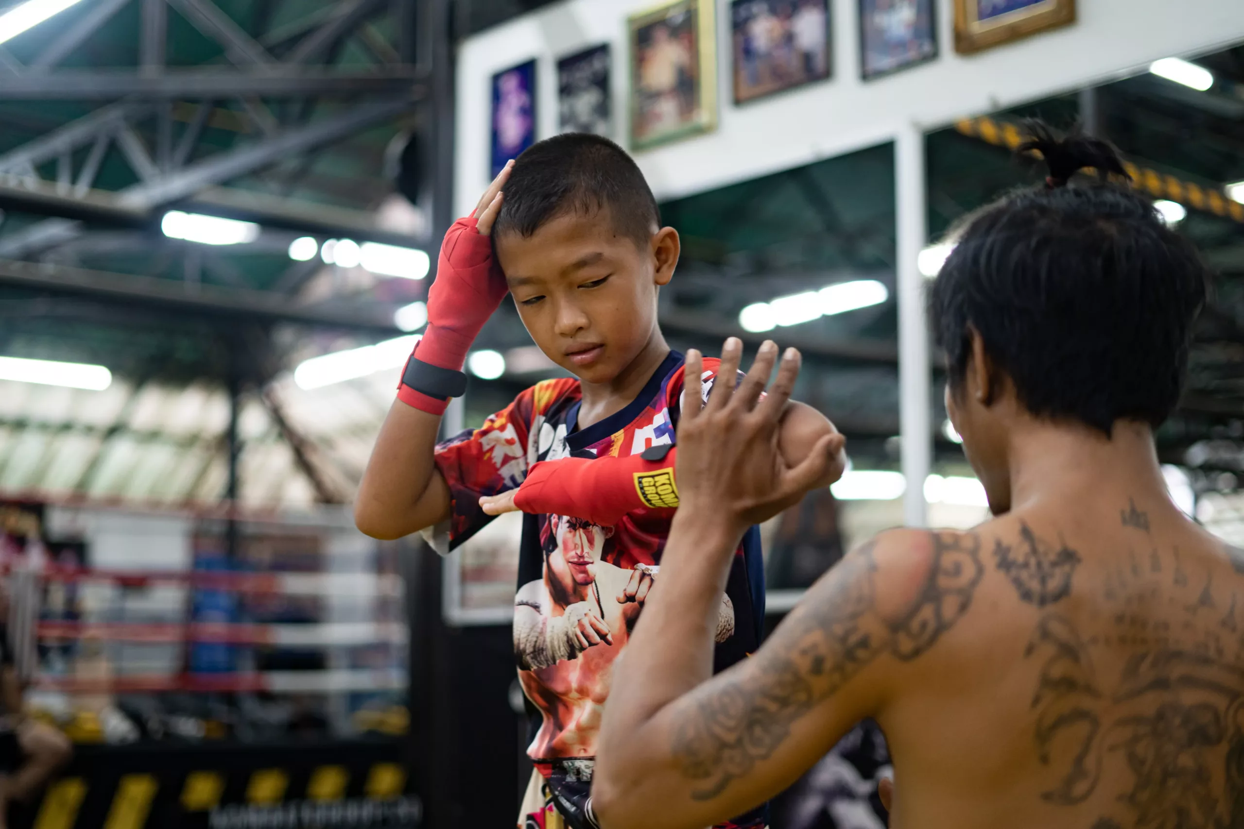 A young boy in Muay Thai attire attentively listens to instructions, wearing hand wraps and a focused expression, as his coach demonstrates a technique in a gym filled with cultural décor.