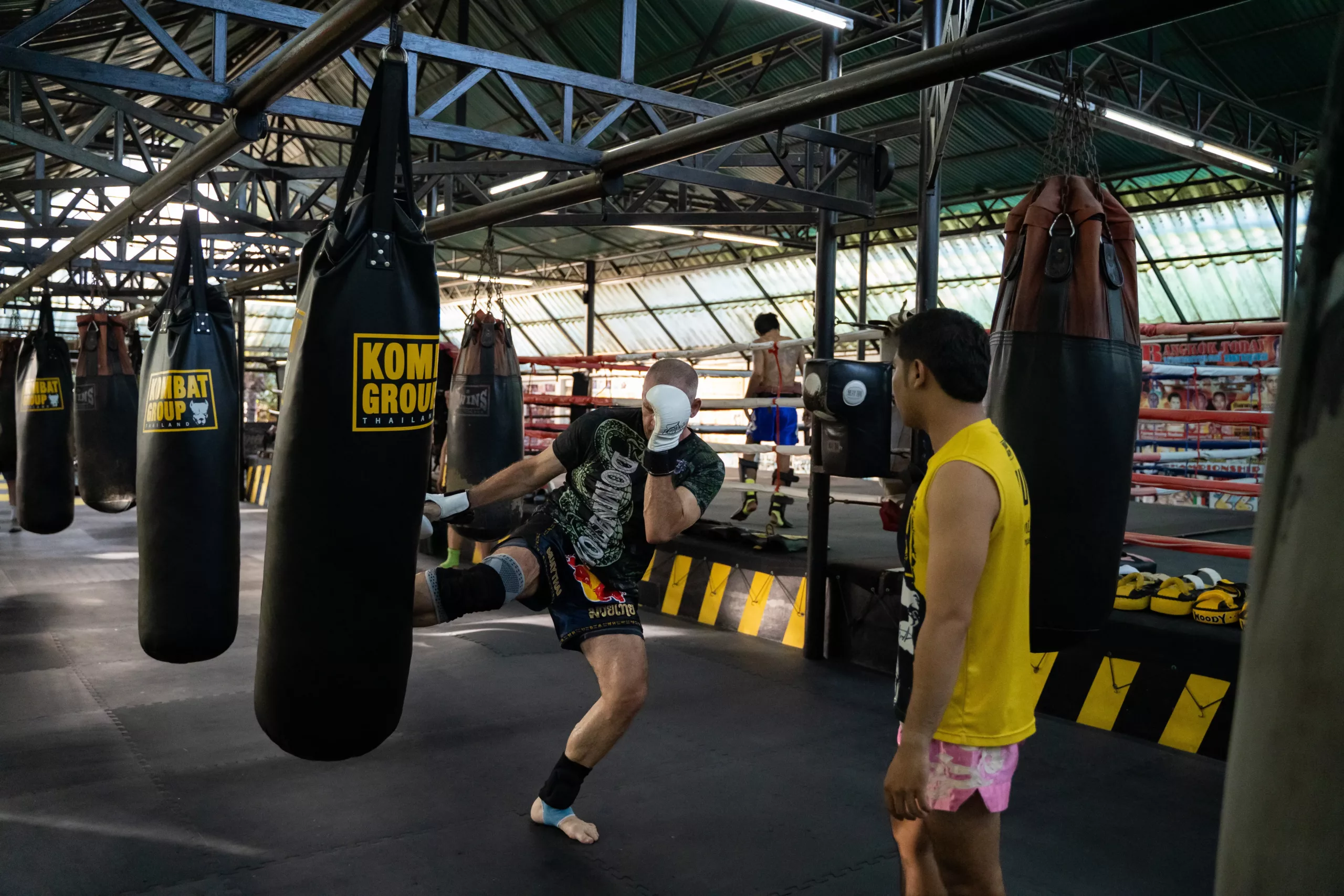 An athlete in Muay Thai shorts is captured in mid-kick against a heavy bag, with a focused trainer observing, in a spacious gym with multiple punching bags and a boxing ring in the background.