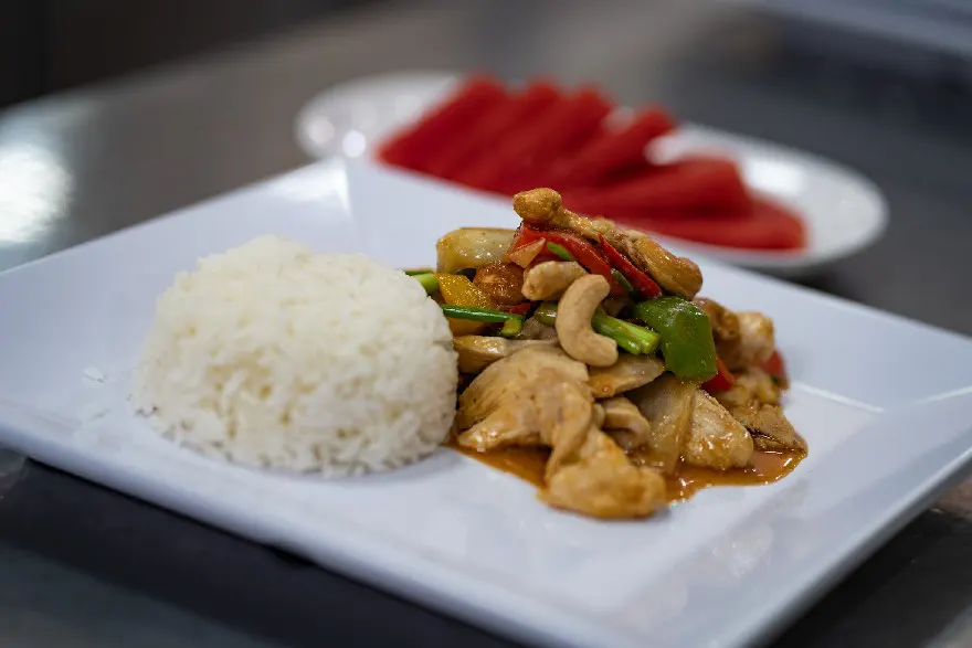 A plate of appetizing cashew chicken paired with a mound of steamed white rice, garnished with green bell peppers and chili, served with a side of sliced watermelon.