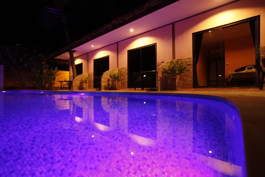 An enchanting evening view of a tranquil swimming pool illuminated with purple lights, reflecting a modern villa with sliding glass doors and cozy interiors, creating a relaxing atmosphere.