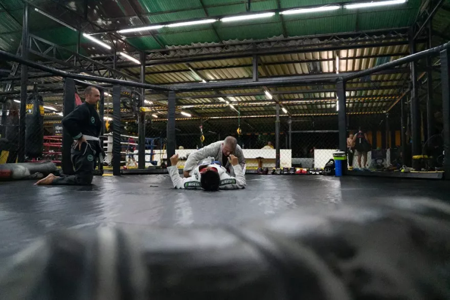 Two Brazilian Jiu-Jitsu practitioners in a sparring session on the mat, with one executing a submission hold as the coach observes closely in a well-equipped martial arts gym.