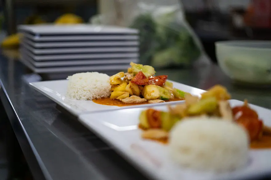 A delectable sweet and sour chicken dish served with a side of fluffy white rice, artistically plated on a white rectangular plate against a professional kitchen backdrop.