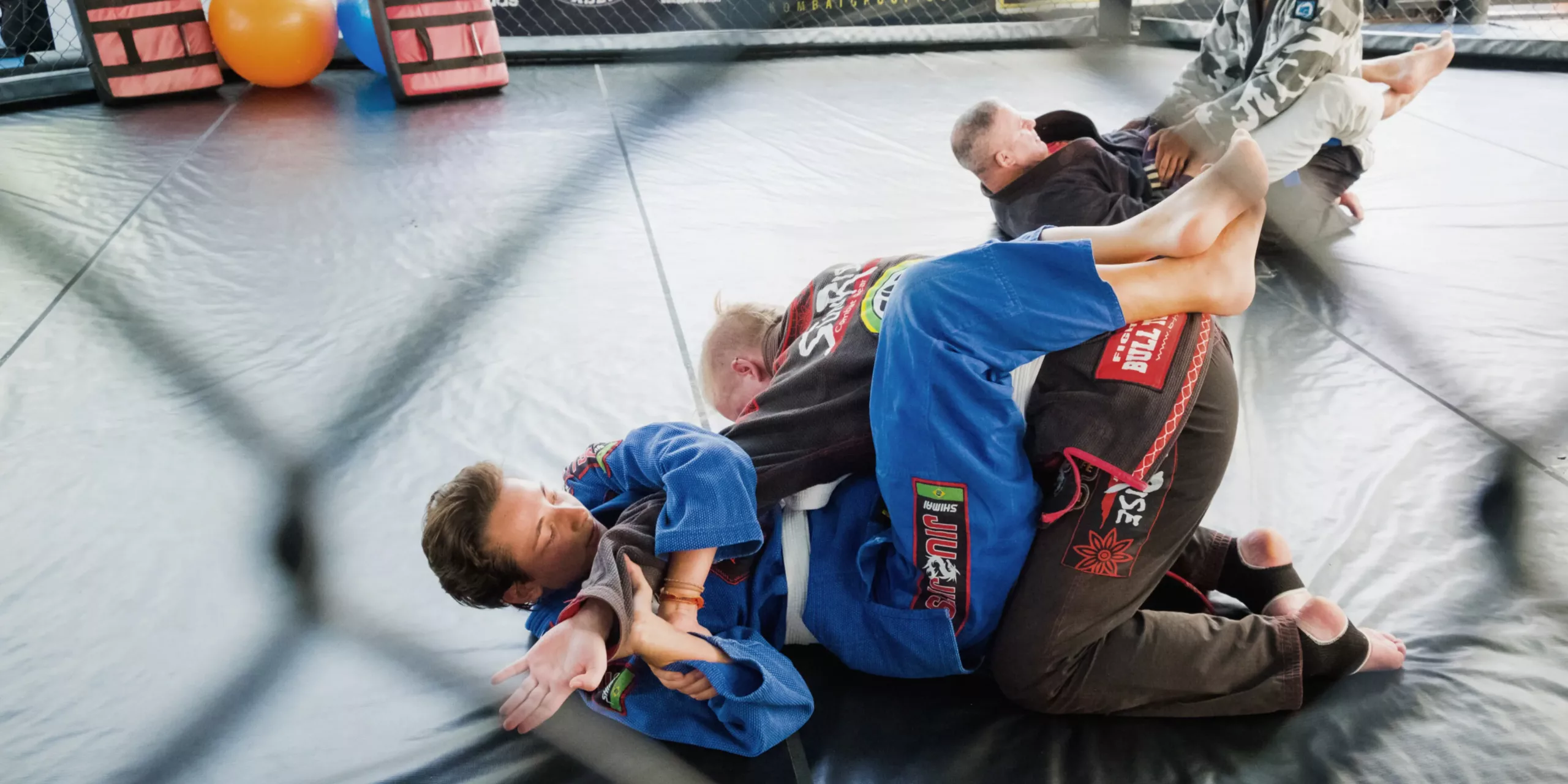 Brazilian Jiu-Jitsu students practice on the mat, with one applying a guard technique while their partner works on passing the guard, showing concentration and teamwork in a grappling drill.