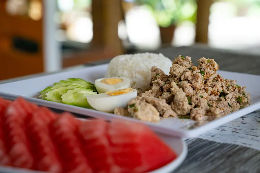 A plate featuring Thai-style minced pork over rice with a hard-boiled egg and cucumber slices, accompanied by fresh watermelon slices, served on a wooden table with a tropical backdrop.