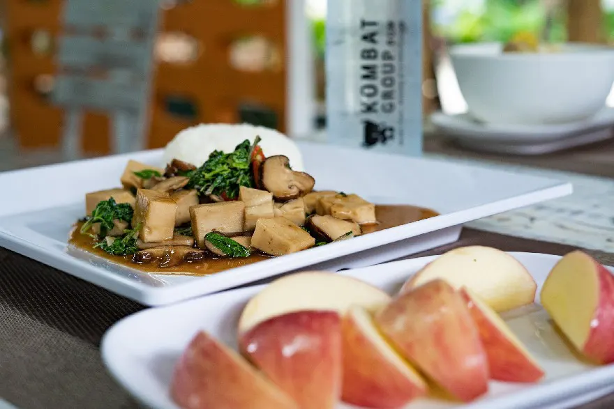 A healthy vegan meal of tofu stir-fry with greens and mushrooms, served alongside white rice and fresh apple slices, with a water bottle in the soft-focus background.