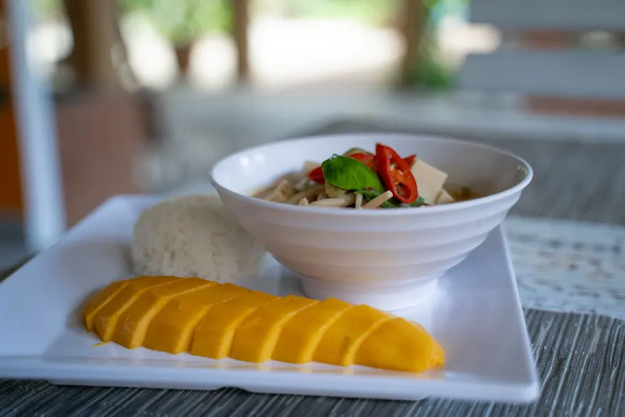 A sumptuous Thai meal featuring ripe mango slices beside a mound of sticky rice, with a bowl of vegetarian mushroom soup garnished with herbs, elegantly served on a white rectangular plate.